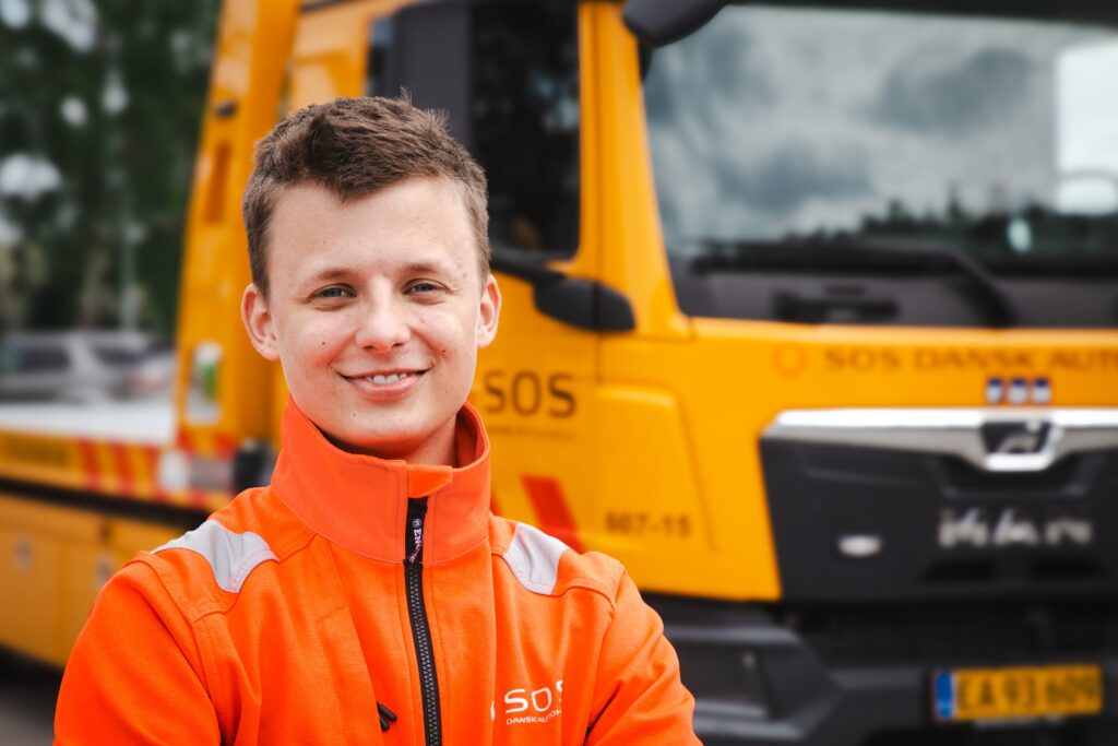 Smilende ung autohjælper i SOS Dansk Autohjælp-uniform foran sin gule bjærgningsbil.